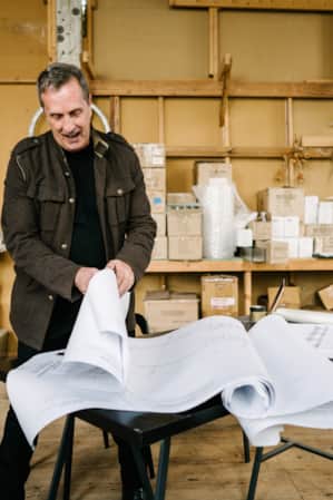 A man standing in a warehouse, thumbing through large pieces of paper with building layout plans.