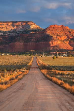 A straight road in the desert leading to a large red rock monument.