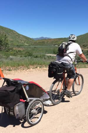 A person biking on a gravel path through green hills, towing a bike trailer.