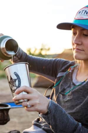 A woman sitting outside in a camp chair, pouring water from a hydro flask into an aluminum drinking cup.