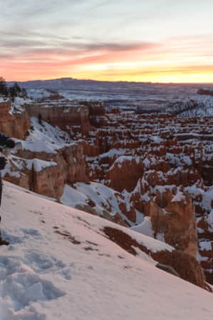 Two hikers overlooking a snowy valley of hoodoos, pouring a warm drink from a thermos.