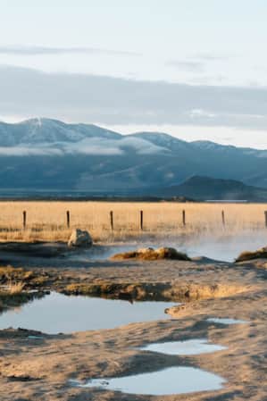 A natural hot spring in a vast field of grass and sand, with a mountain range visible in the distance.