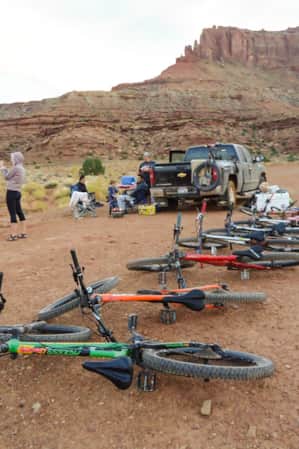Mountain bikes lying on their sides in a row in the desert, with a group of people by a truck with camping gear off to the side.