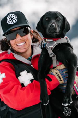 A ski patrol employee smiling while holding a black Labrador Retriever.