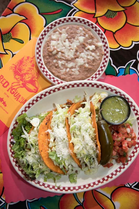 Three hard-shell corn tortillas served with shredded lettuce, tomatoes, sauce and refried beans from The Red Iguana.