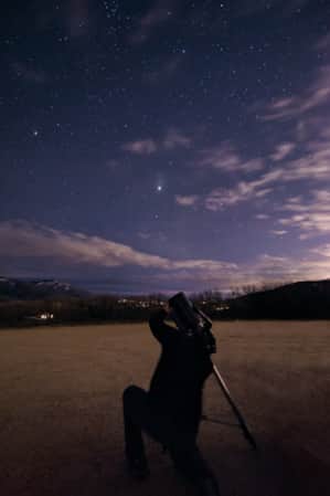 A person kneeling and looking through a telescope at a star-filled night sky.