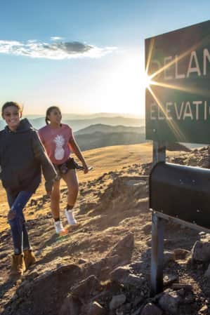 Two young girls standing at the summit of a mountain next to a sign that reads 'Delano Peak Elevation 12,169'.