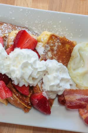 Aerial view of a plate with french toast topped with whipped cream and strawberries, with a fried egg and bacon on the side.