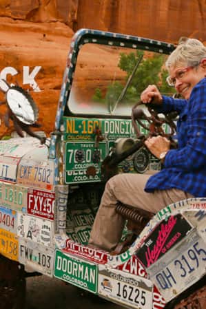 A woman pretending to drive a car made out of license plates with wrought-iron wheels.