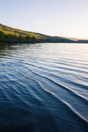 Landscape view of a lake with green mountains covered in trees and bushes on the left side, and the sun gleaming over the top of a mountain on the horizon to the right.