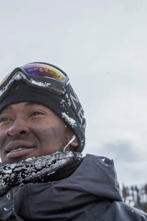 A man standing outdoors with mountains in the background, smiling as he gazes into the distance, dressed warmly in winter clothes.