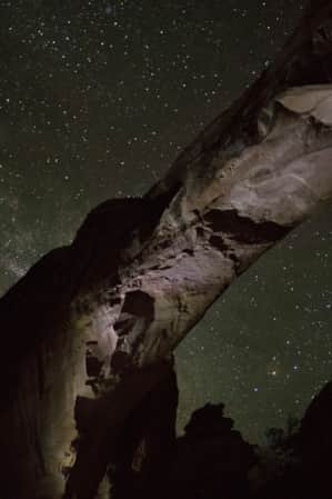 The underside view of a natural rock arch against a starry night sky.