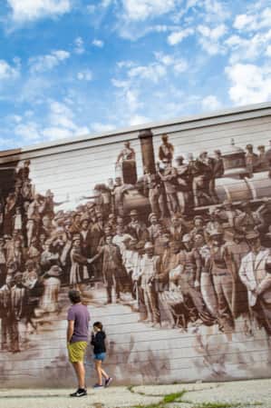A man and a young girl admiring a large mural depicting an old-time scene of people at a railroad.