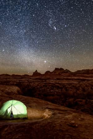 An expansive view of a red rock desert illuminated by the night sky, with a green tent in the landscape and a light on inside the tent.