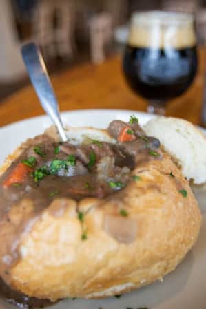 A bread bowl filled with beef stew, accompanied by a dark beer on the side.