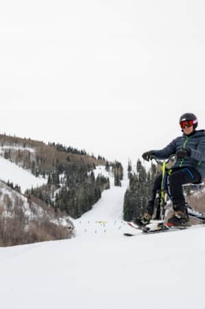 Two people skiing down a mountain: one on special assisted skis, with a man attached by a rope riding behind.