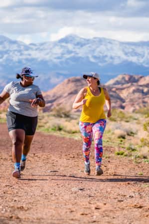 Two women running on a dirt trail, surrounded by open fields, with a mountain range visible in the background.