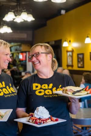 Two women smiling and looking at each other while standing in a restaurant, each holding a plate of breakfast foods.