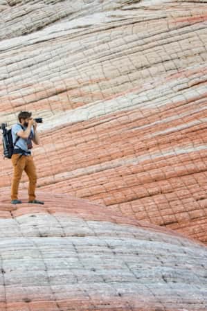 A man with a large camera taking a photo of something in the distance, standing on and surrounded by large rock formations with white and red striations.