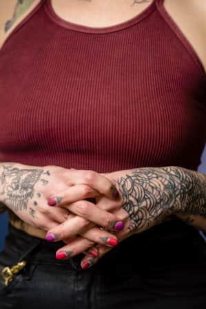 A torso shot of a woman with her fingers intertwined near her stomach, wearing a tank top and showing tattoos on both arms.