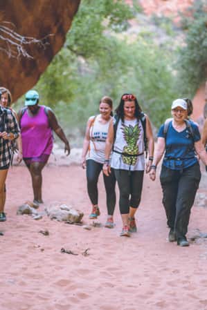 A group of women hiking on sand through the desert, with large red rocks on either side.