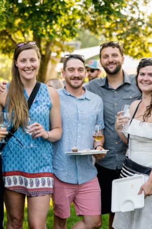 People standing next to each other, all smiling, with some holding wine glasses, at a festival, with others walking around in the background.