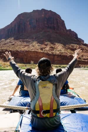 Three people on a large raft in a river, with one person holding their hands in the air, passing by a red rock monument.