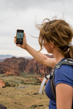 A woman hiking with a backpack, holding up her phone to take a picture of a landscape with sparse vegetation and rock formations.