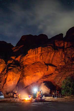 People sitting around a campsite at night, surrounded by red rock formations, with the area illuminated by the light of a campfire.