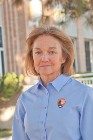 A woman standing in front of a brick building, wearing a blue shirt with the National Park Service logo.