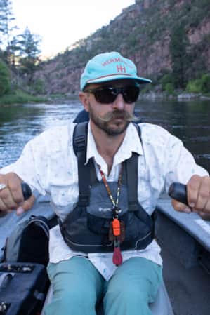 A man rowing a canoe in a river, with fly fishing gear visible in the canoe.
