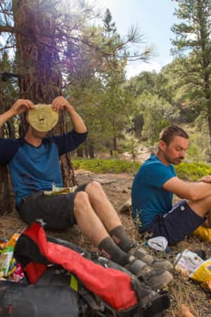 Two mountain bikers resting under a tree, eating a snack.