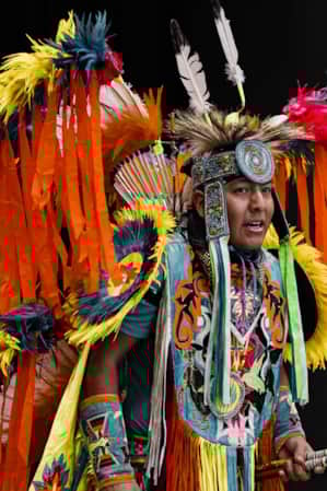 A man dressed in traditional Navajo Hoop dancing attire, featuring orange and yellow feather wings, a feathered headpiece, and a turquoise chest covering with traditional patterns.