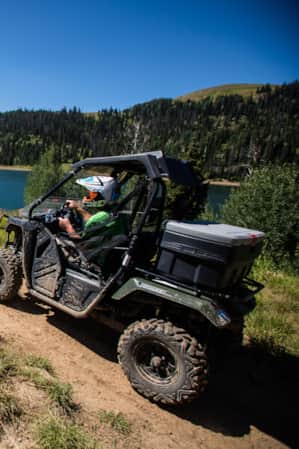 Two ATVs drive along a dirt path beside a lake.