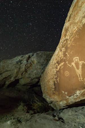 Petroglyphs on a large rock illuminated by soft light, set against a night sky filled with bright stars.