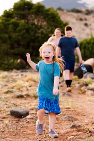 A young girl running on a hiking trail, smiling with her hand up in the air, while her family walks behind her.