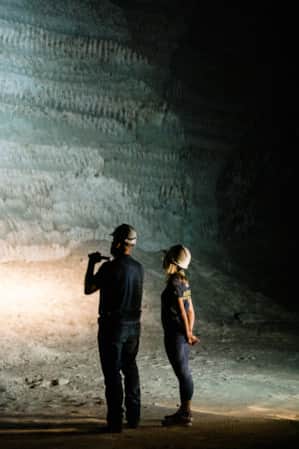 A man and a woman wearing hard hats and holding flashlights, standing next to each other at night, looking at a tall rock slab with various formations.