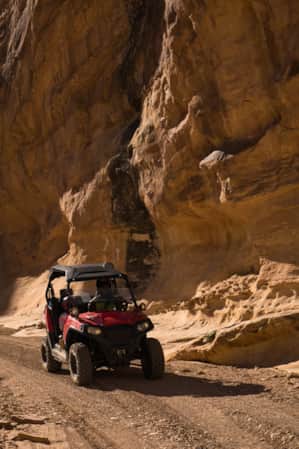 A person riding an ATV along a sandy trail, with greenery on one side and a large rock formation providing shade on the other.