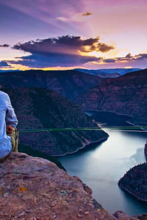 A person holding a fishing rod, overlooking a canyon with a river at the base.