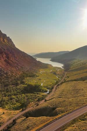 A large red rocky and sandy mountain stands on the left side of the landscape, with a road winding through a lush green valley. A wide stream flows into a narrow river at the mountain’s base.