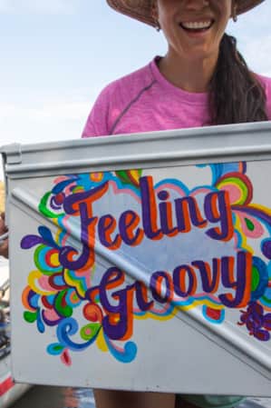 A woman smiling and holding a handwritten sign that reads 'feeling groovy,' with a raft visible behind her at a lake.