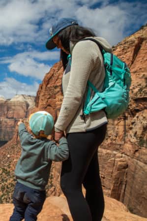 A woman and her child overlooking a vast canyon with impressive rock formations and a winding road below.