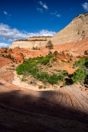 A desert landscape with vibrant red and orange sandstone formations, featuring layered rock patterns and sparse green vegetation under a clear blue sky.