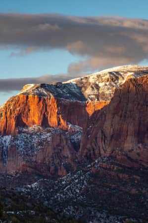 Large red rock mountains with a light dusting of snow, and their peaks illuminated by a ray of sunshine.