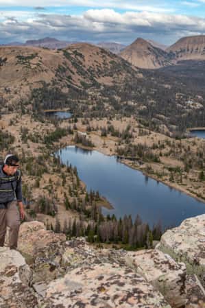 A man hiking at the top of a mountain, overlooking multiple lakes scattered across the base and additional mountain ranges in the background.