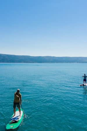 Two men stand-up paddleboarding on a lake with mountains in the backdrop.