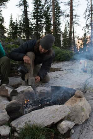 A man and a woman drinking warm beverages around a fire pit in a forest.