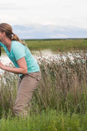 A woman and a young child, standing in the grass by a river, pointing and looking through binoculars at something in the distance.