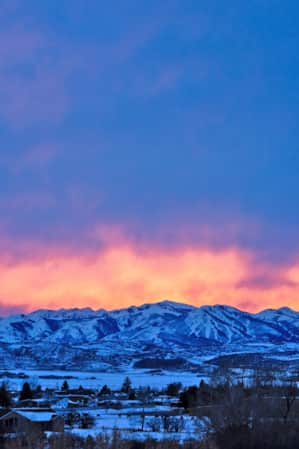 A range of snow-capped mountains set behind a spacious neighborhood at sunset, with deep oranges and pinks filling the sky.