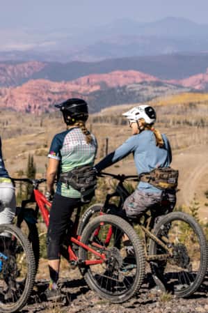 Three women resting on their mountain bikes, overlooking a valley with mountains in the distance.